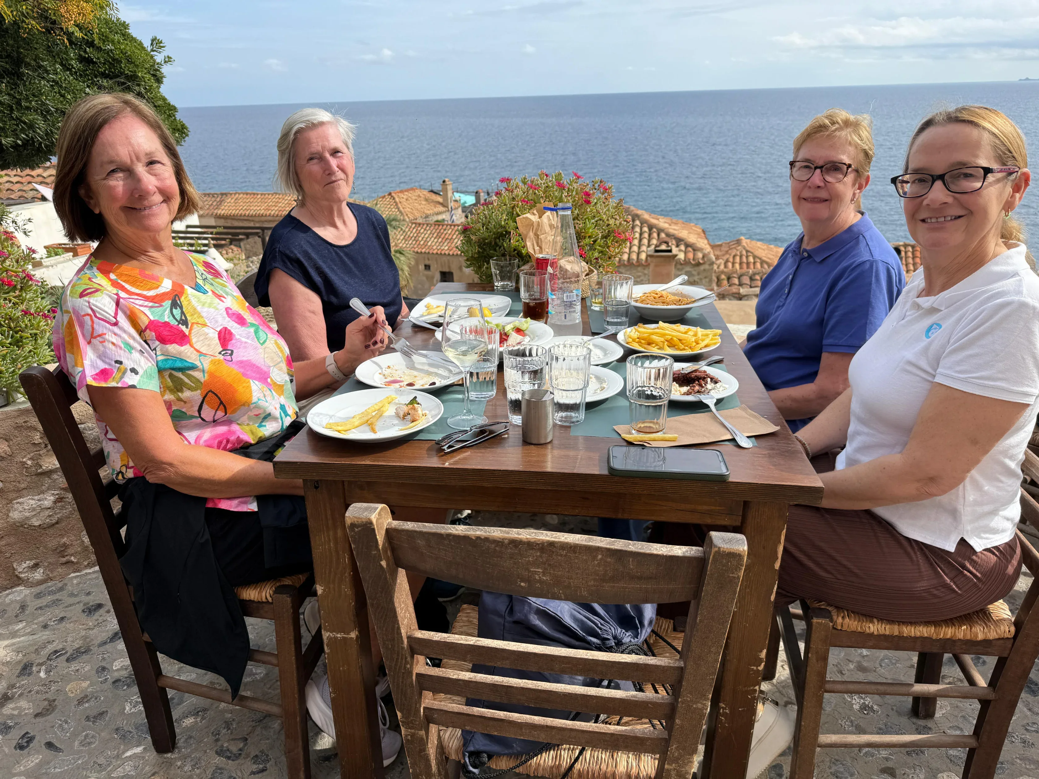 Four women sit at an outdoor table with finished plates and drinks, overlooking rooftops and the sea under a partly cloudy sky.