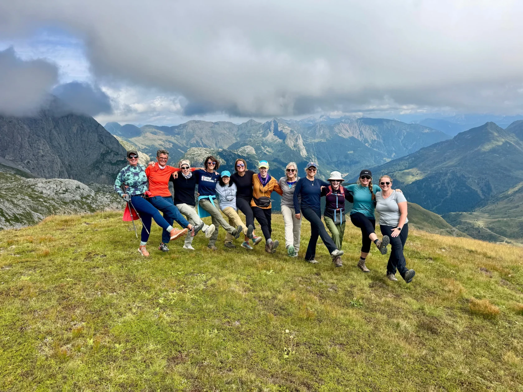 A group of eleven people stand in a line on a grassy hill, linking arms and lifting one leg, with mountains and clouds in the background.