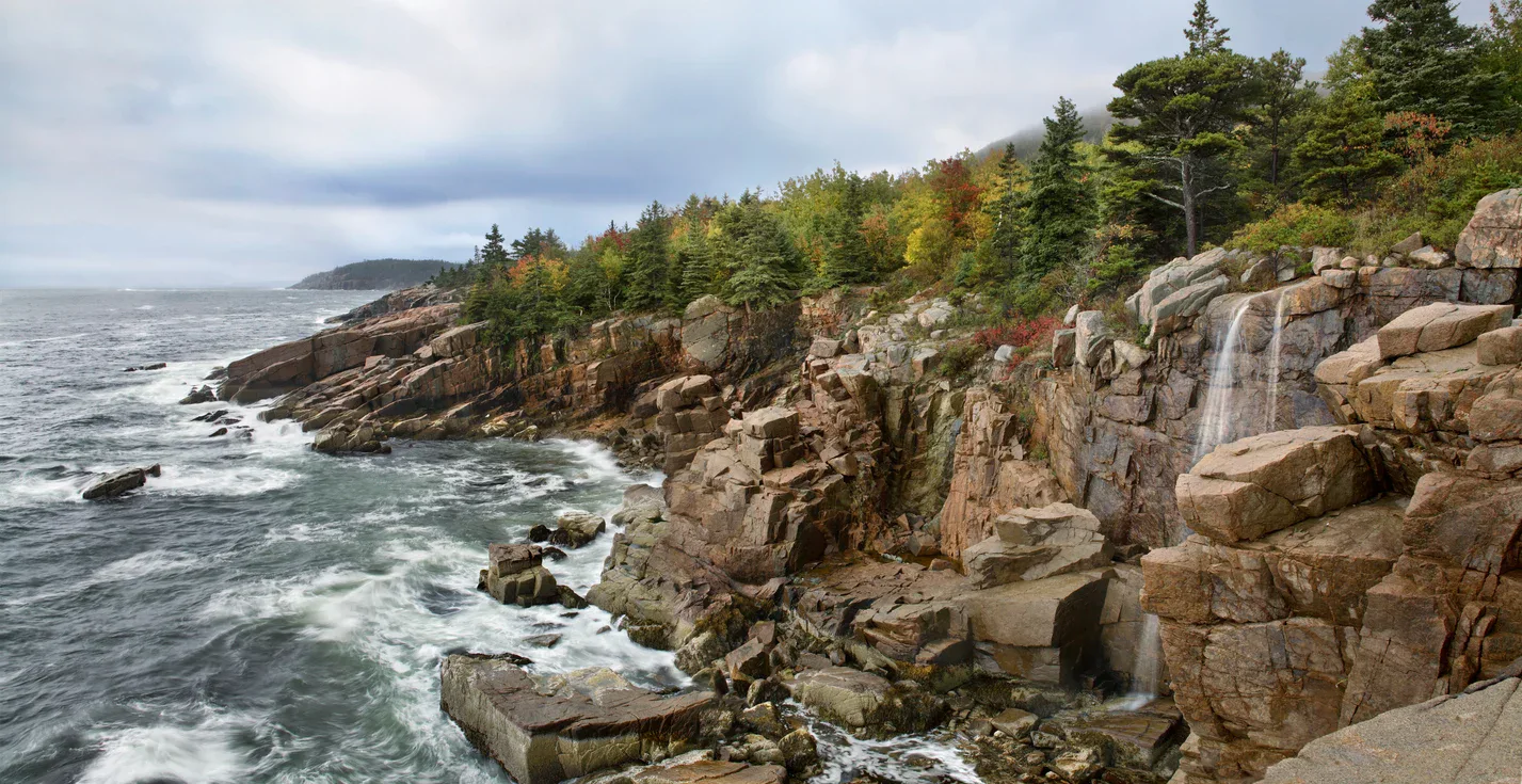 Rocky coastline with waves crashing against large boulders, a small waterfall, and a forest of evergreen and autumn-colored trees under a cloudy sky.