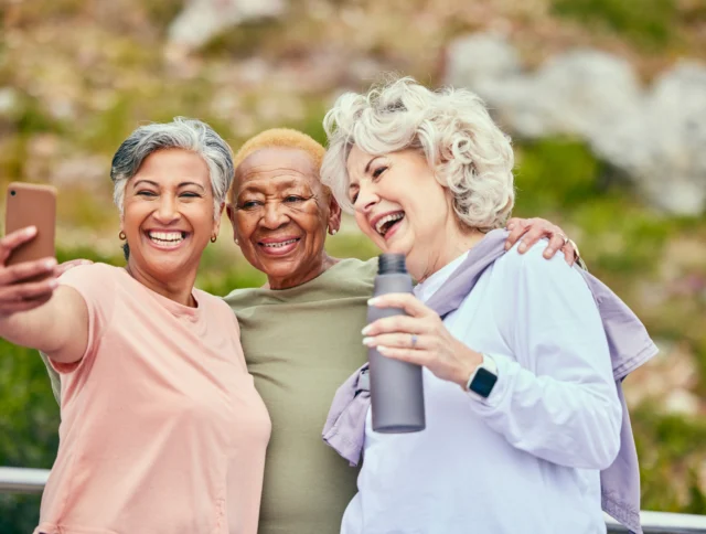 Three older women smiling and posing for a selfie outdoors, with one holding a phone and another holding a water bottle.