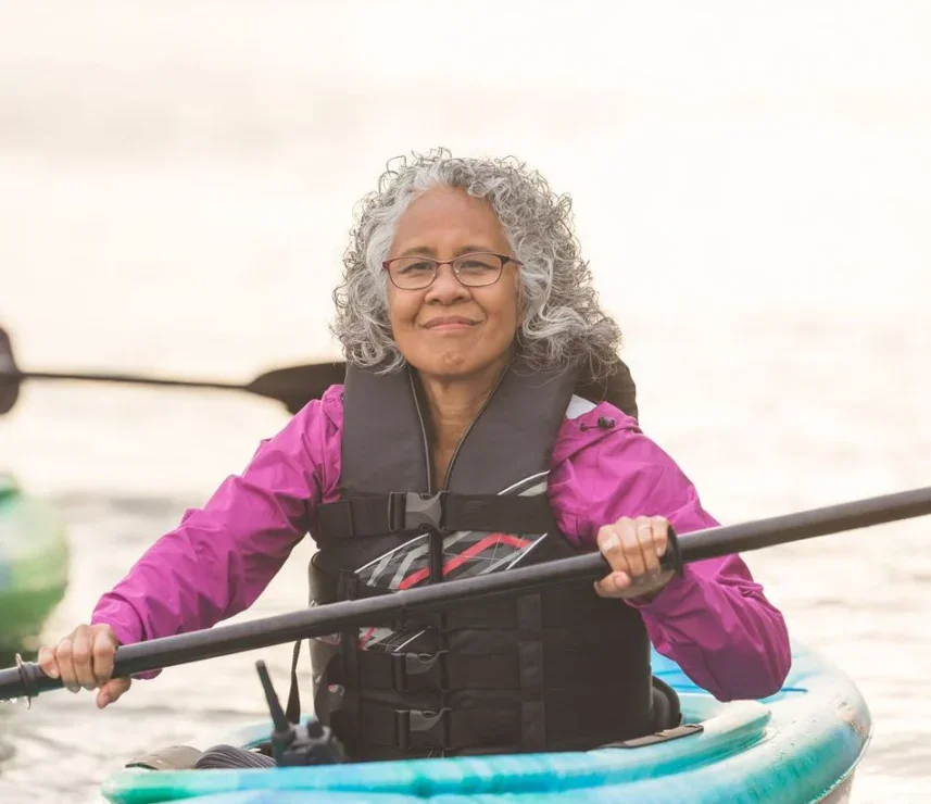 An older woman with curly gray hair wearing glasses and a life jacket paddles a kayak on calm water.
