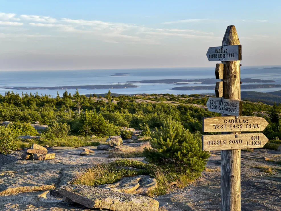 Wooden trail signpost stands on a rocky hilltop with green trees and distant ocean islands visible under a clear blue sky.