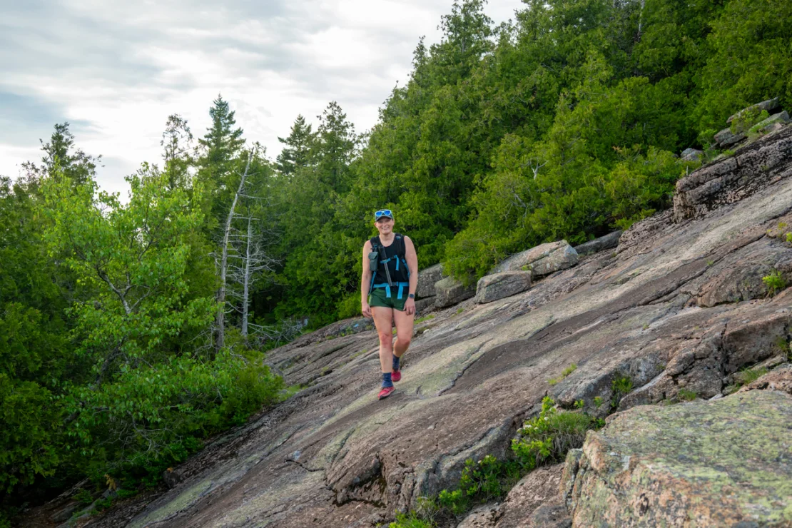 A person wearing athletic gear and a backpack walks on a sloped rocky terrain surrounded by green trees under a cloudy sky.