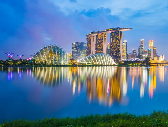 Singapore skyline at dusk featuring Marina Bay Sands, Supertree Grove, and glass domes of Gardens by the Bay, reflected in calm waterfront.