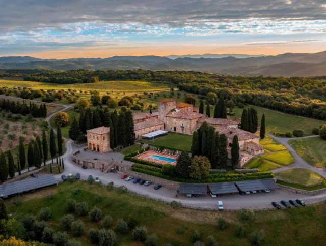 Aerial view of a large Tuscan villa with terracotta roofs, surrounded by trees, gardens, parked cars, and rolling hills under a partly cloudy sky at sunset.