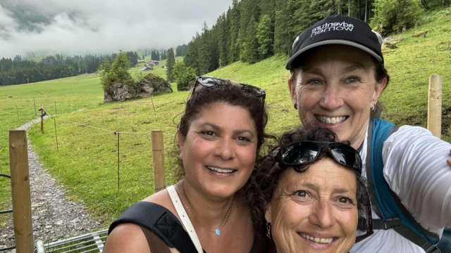 Three women smiling for a selfie outdoors on a cloudy day, standing on a trail with green grass, trees, and hills in the background.