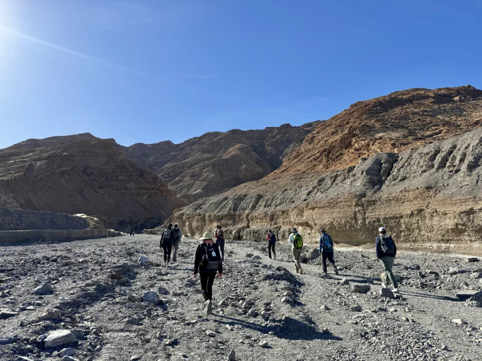 A group of people with backpacks hike through a rocky canyon under a clear blue sky, surrounded by layered hills and dry terrain.