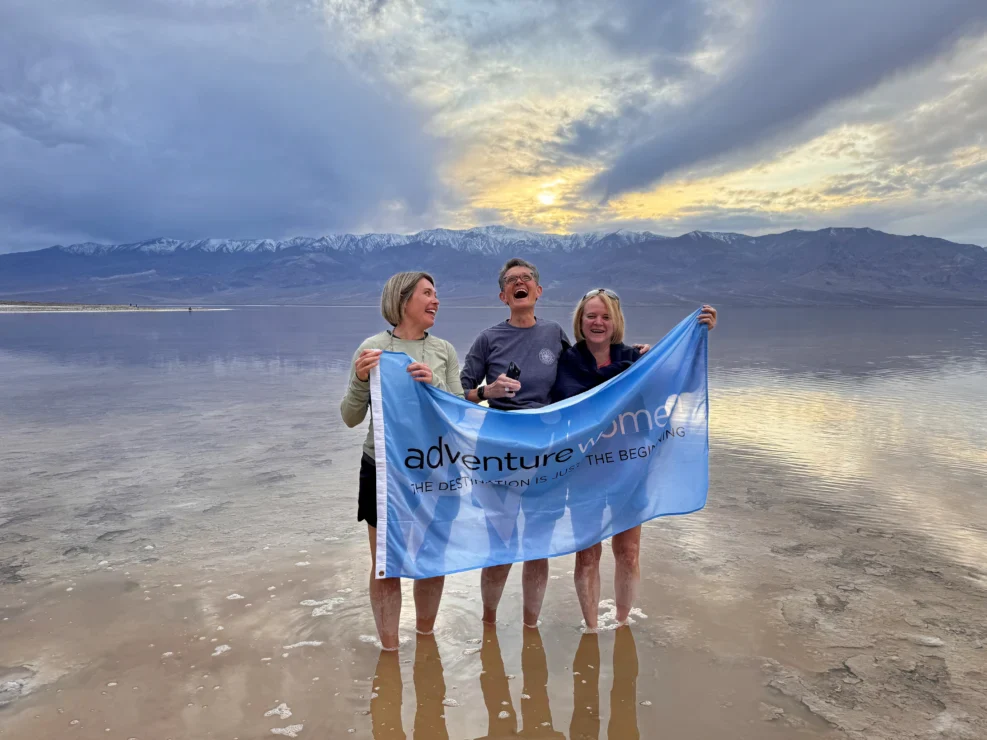 Three people stand barefoot in shallow water holding an “AdventureWomen” banner, with mountains and a cloudy sunset sky in the background.