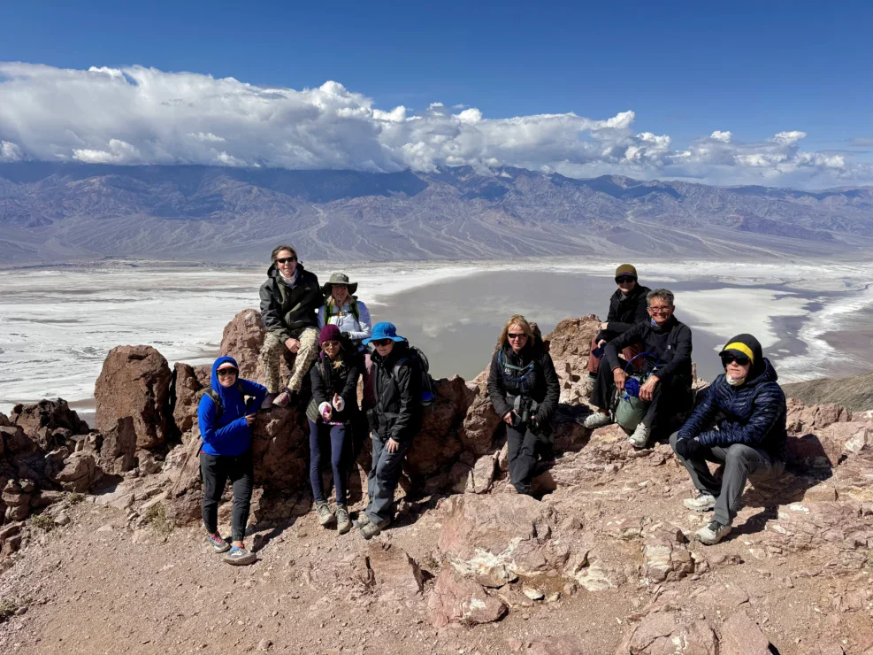 Nine people sit and stand on rocky terrain overlooking a desert valley and mountains under a blue sky with clouds.
