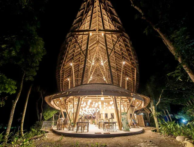 A bamboo structure with a tall, conical roof is illuminated at night, with chairs and tables arranged inside and trees surrounding the building.