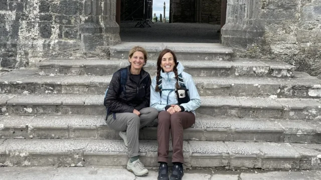 Two people sit on stone steps in front of a large arched doorway in a weathered stone building, with another archway and a bright opening visible in the background.