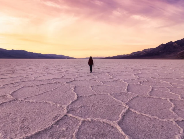 A person walks alone across a cracked, salt flat landscape at sunset with mountains visible in the background.