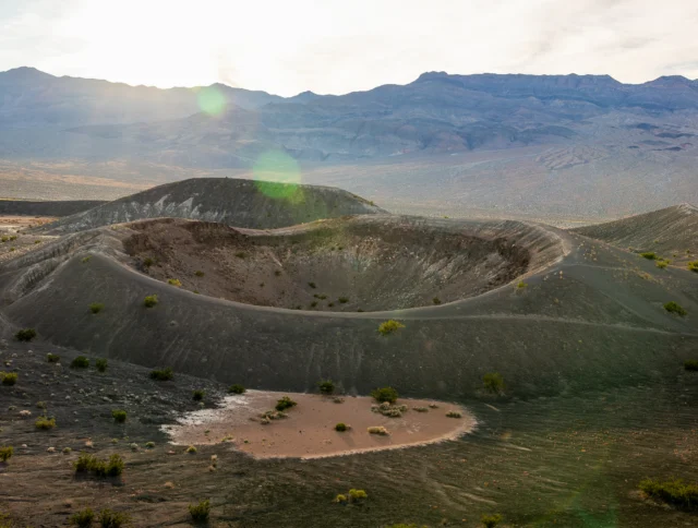 Large volcanic crater with sparse vegetation in a desert landscape, mountains in the background, and sun shining from the upper left.