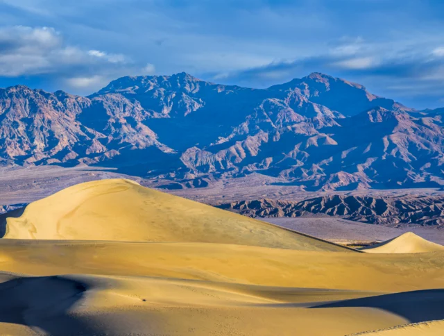 Golden sand dunes in the foreground with rugged, rocky mountains and a partly cloudy blue sky in the background.