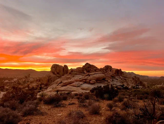 A rocky desert landscape with scattered shrubs at sunset, featuring a dramatic sky with orange, pink, and purple clouds.
