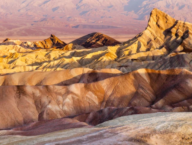 Eroded, multicolored badlands and rocky hills are shown under warm sunlight at Zabriskie Point in Death Valley National Park.