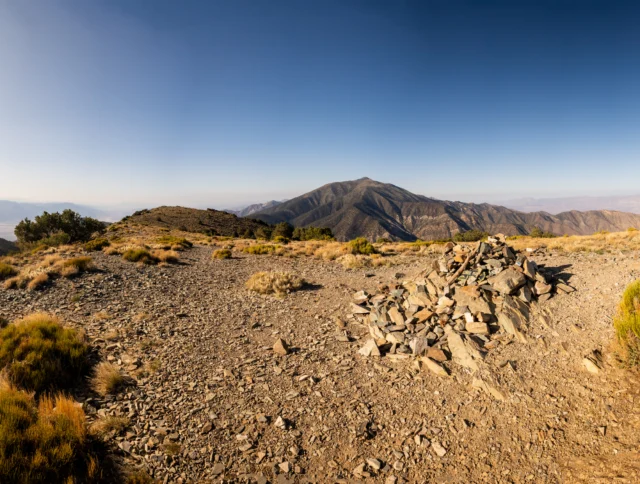 Rocky mountain landscape with sparse vegetation, a pile of stones in the foreground, and distant peaks under a clear blue sky.