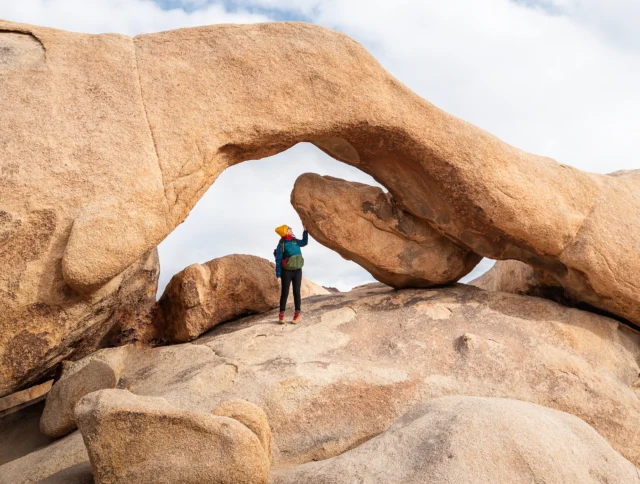 A person wearing a yellow hat stands on large rocks, touching a natural rock arch formation under a cloudy sky.
