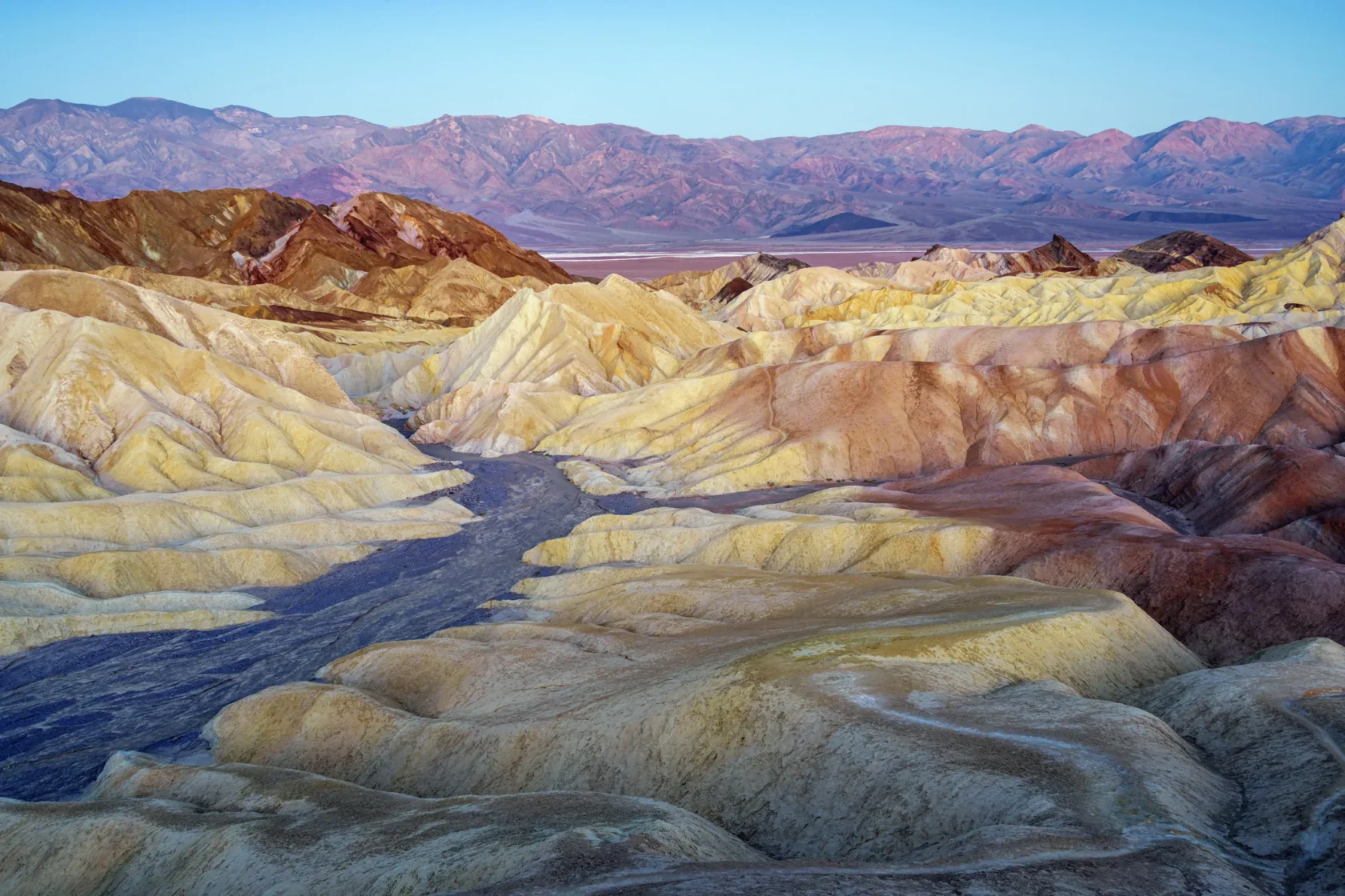 Colorful, eroded hills and ridges under a clear sky at Zabriskie Point in Death Valley National Park, with distant mountains in the background.