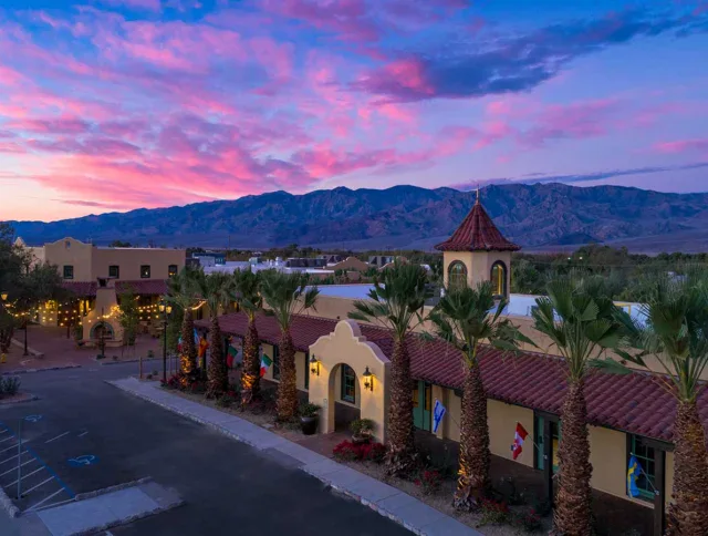 A southwestern-style building with palm trees in front, lit by string lights at dusk, with mountains and a colorful sky in the background.