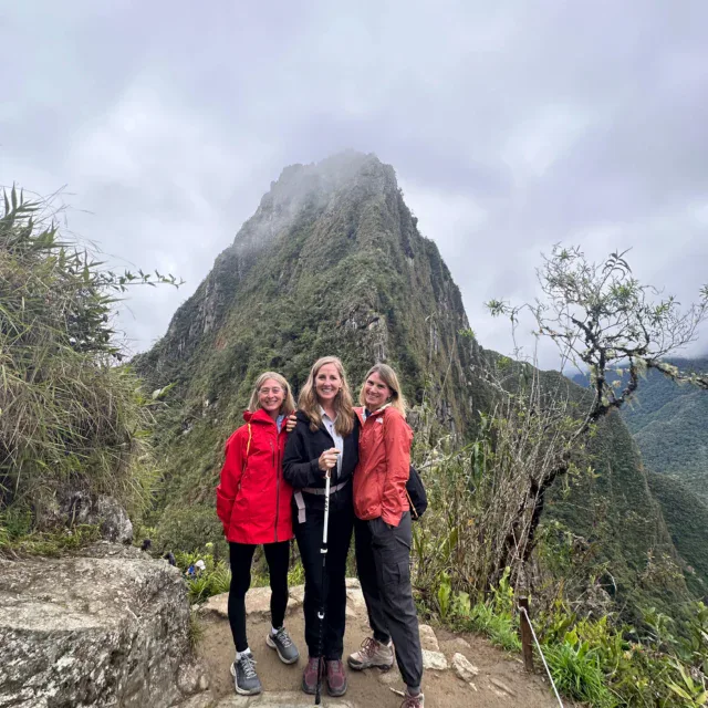 Three people pose on a rocky trail with a lush, steep mountain peak and cloudy sky in the background.