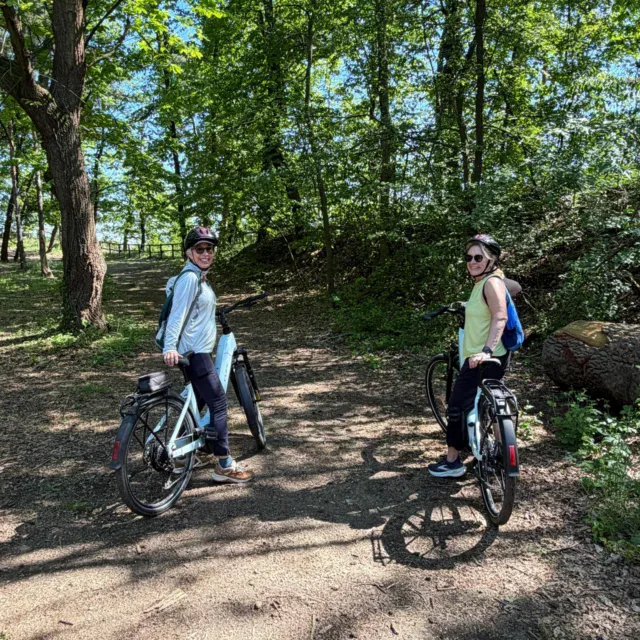 Two people wearing helmets stand with bicycles on a dirt path in a wooded area, looking back toward the camera under bright sunlight.