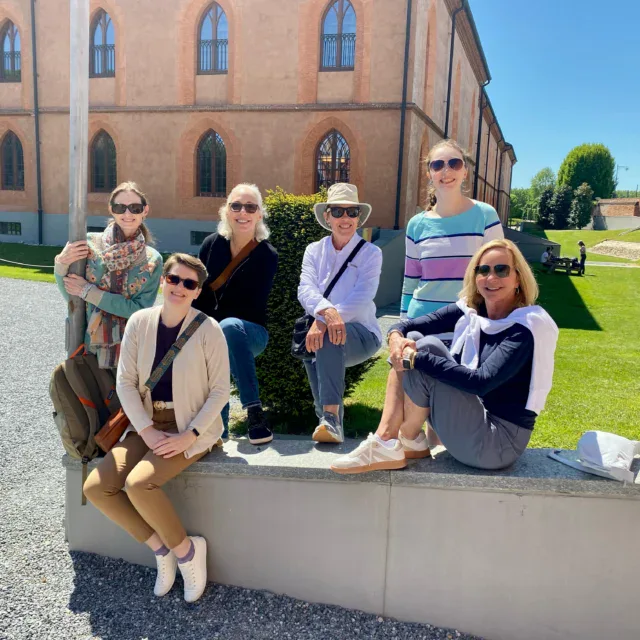 Six adults pose and smile for a group photo outdoors on a sunny day, seated and standing by a low wall in front of a large historic building with arched windows.