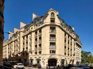 A large, cream-colored stone building with multiple floors, arched windows, and French flags, situated on a street corner under a clear blue sky.