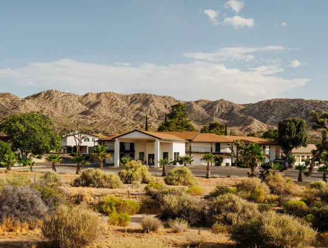 A large, white, two-story building with a brown roof stands amid desert vegetation, with mountains and a partly cloudy sky in the background.