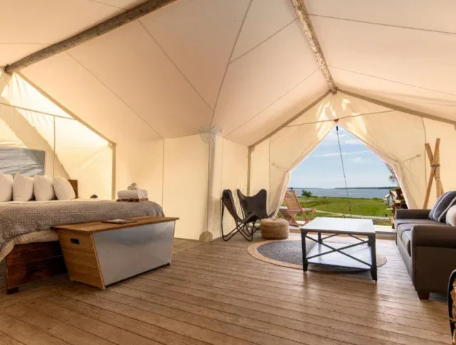 Spacious glamping tent interior with a bed, couch, coffee table, and wooden flooring, overlooking a grassy area and water through an open entrance.