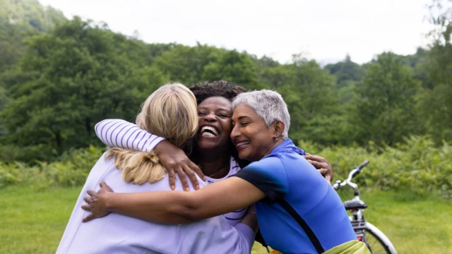 Three women hug each other and smile outdoors on a grassy area, with trees and a bicycle visible in the background.