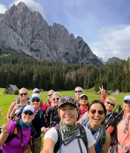 A group of hikers pose and smile in a grassy field with backpacks, with mountains and trees in the background under a partly cloudy sky.