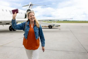A woman stands on an airport tarmac, smiling and holding up her passport and boarding pass near a parked airplane.