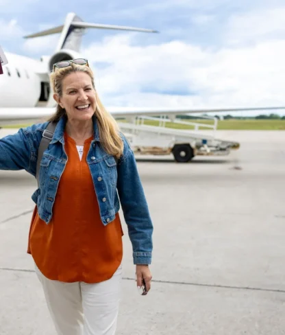 A woman stands on an airport tarmac, smiling and holding up her passport and boarding pass near a parked airplane.