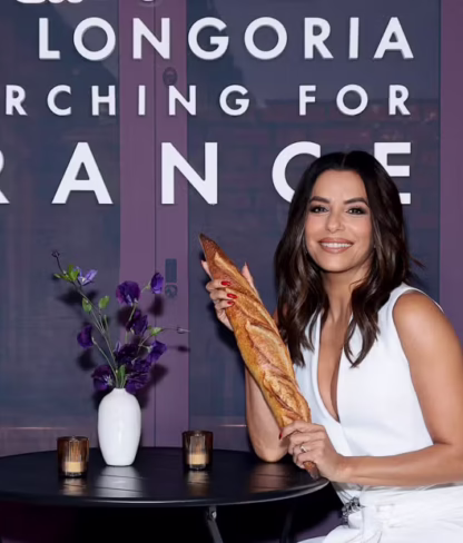 A woman in a white outfit sits at a small round table holding a baguette, with flowers and candles on the table, in front of a sign that reads "Searching for France.