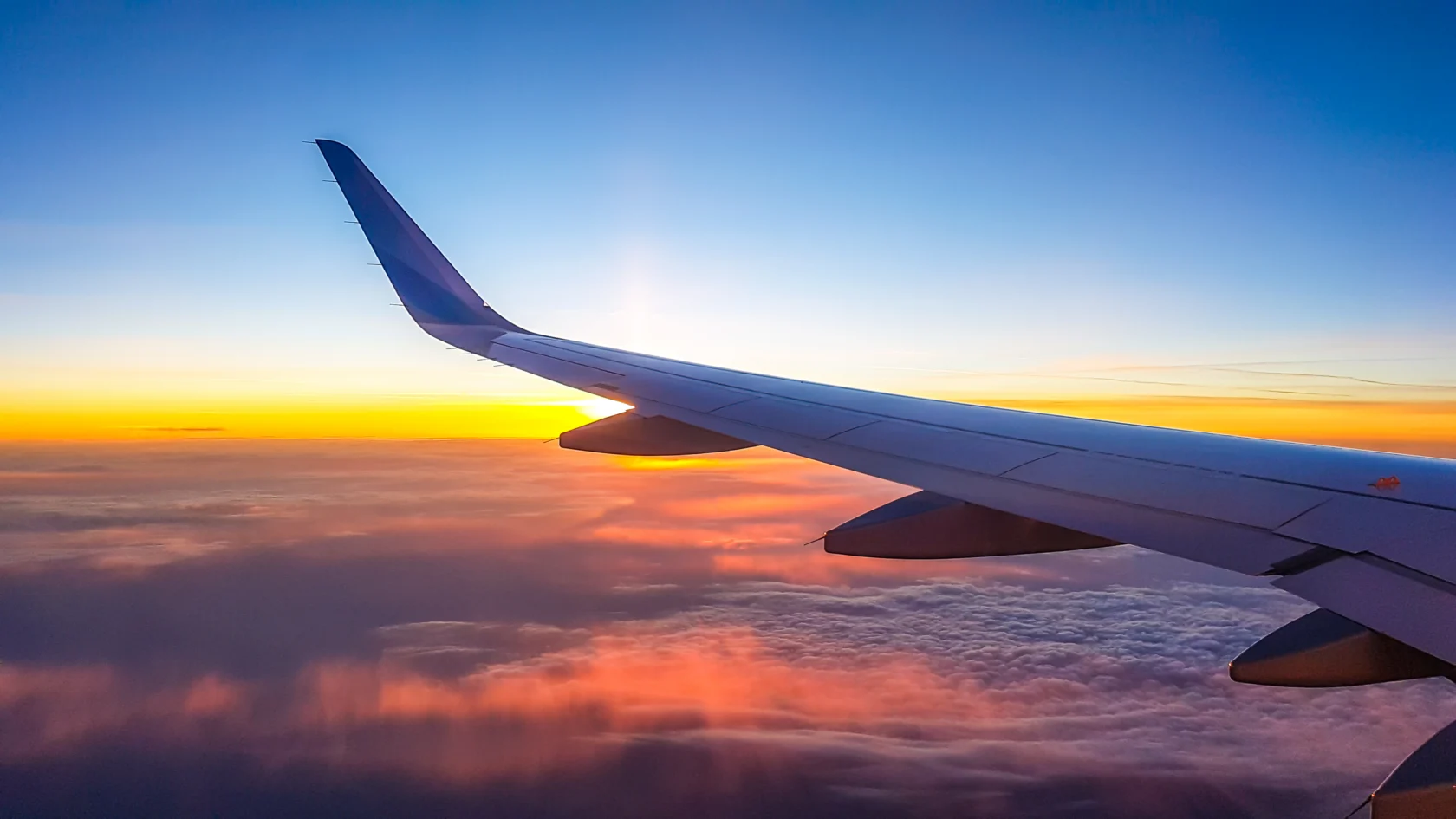 View of an airplane wing above the clouds during a vibrant sunset, with orange and blue hues in the sky.