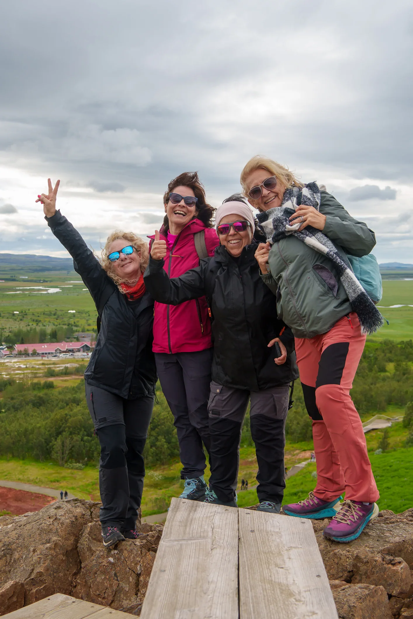 Four women in outdoor clothing stand together on a rocky hill, smiling and posing for the camera with a green landscape and cloudy sky in the background.