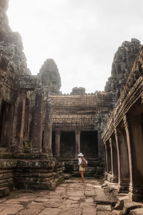 A person wearing a white hat and backpack walks through ancient stone ruins with tall, weathered pillars and walls under a cloudy sky.