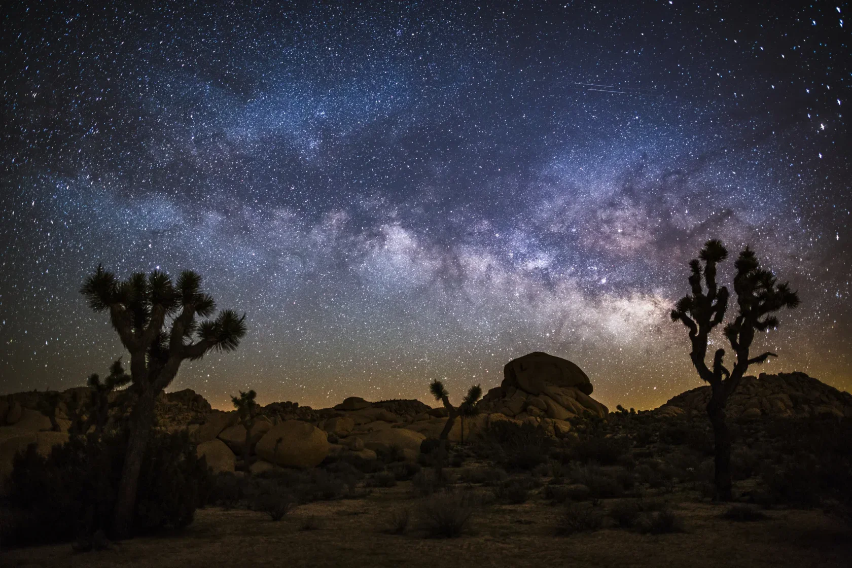Star-filled night sky with the Milky Way visible over a desert landscape featuring Joshua trees and large rock formations.