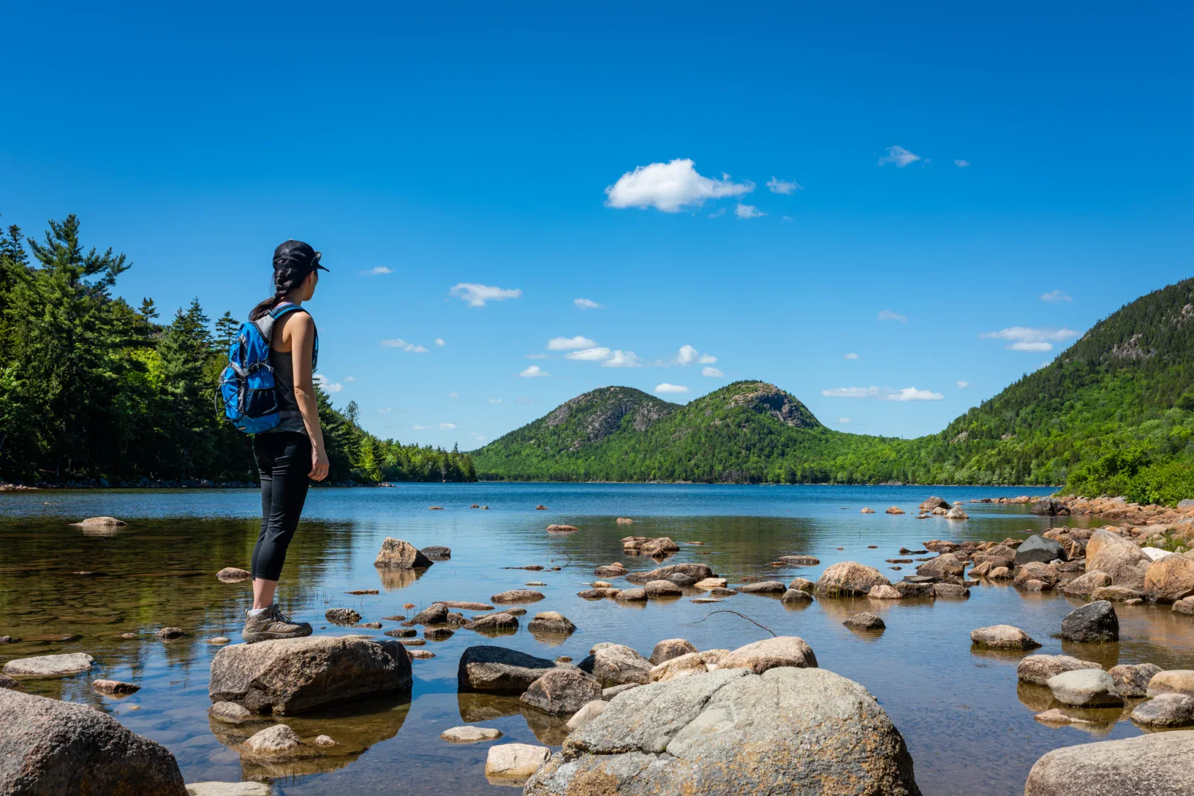 A person with a backpack stands on rocks by a calm lake, looking toward forested hills under a clear blue sky.