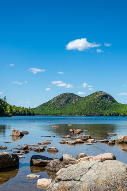 A person with a backpack stands on rocks by a calm lake, looking toward forested hills under a clear blue sky.