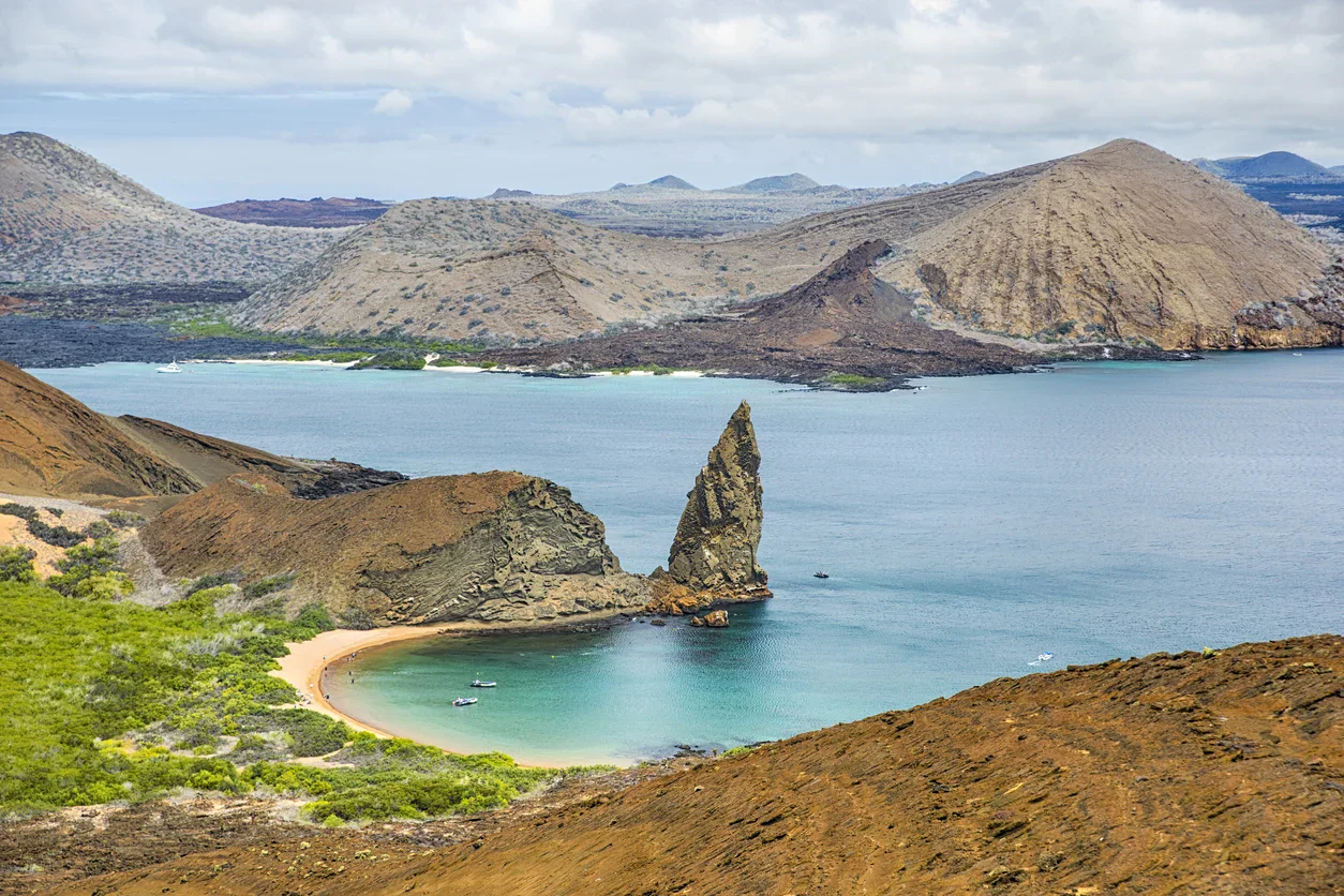 A rocky spire rises from the blue water at a bay surrounded by volcanic hills and sparse vegetation under a partly cloudy sky.