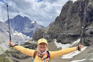 A person in hiking gear stands smiling with arms raised, holding trekking poles, in a rocky mountain landscape with patches of snow under a cloudy sky.