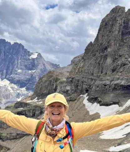 A person in hiking gear stands smiling with arms raised, holding trekking poles, in a rocky mountain landscape with patches of snow under a cloudy sky.