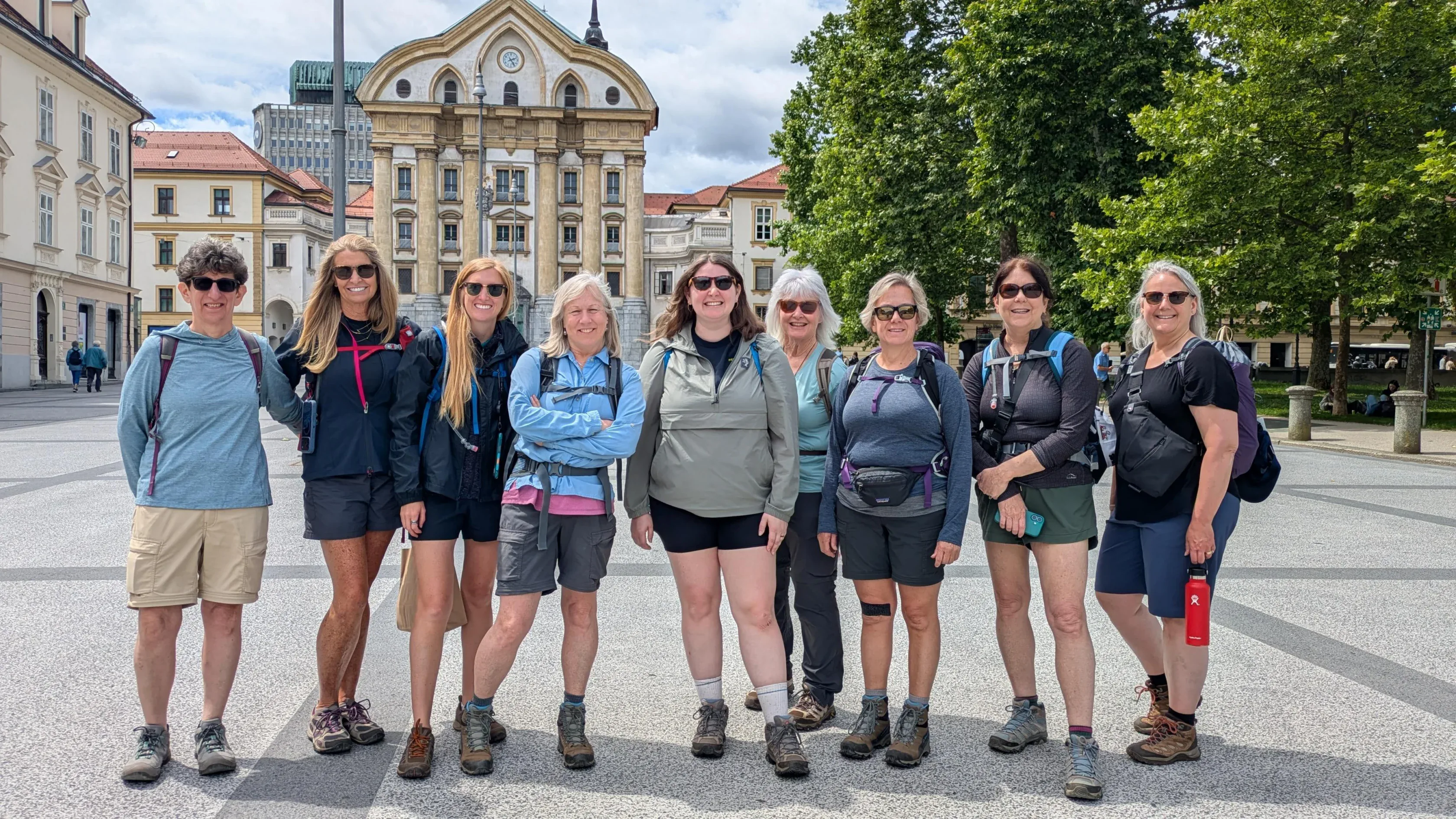 Nine women in casual outdoor clothing stand together in a city square with historic buildings and trees in the background on a sunny day.