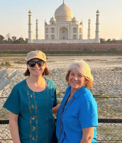 Two women in blue tops stand in front of the Taj Mahal on a sunny day, smiling at the camera.