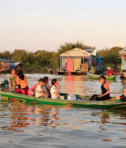 A small wooden boat with several people aboard moves across a calm river, with colorful floating houses and greenery visible in the background.
